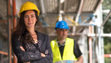 Woman stands wearing a yellow construction hat with a man in PPE on a construction site